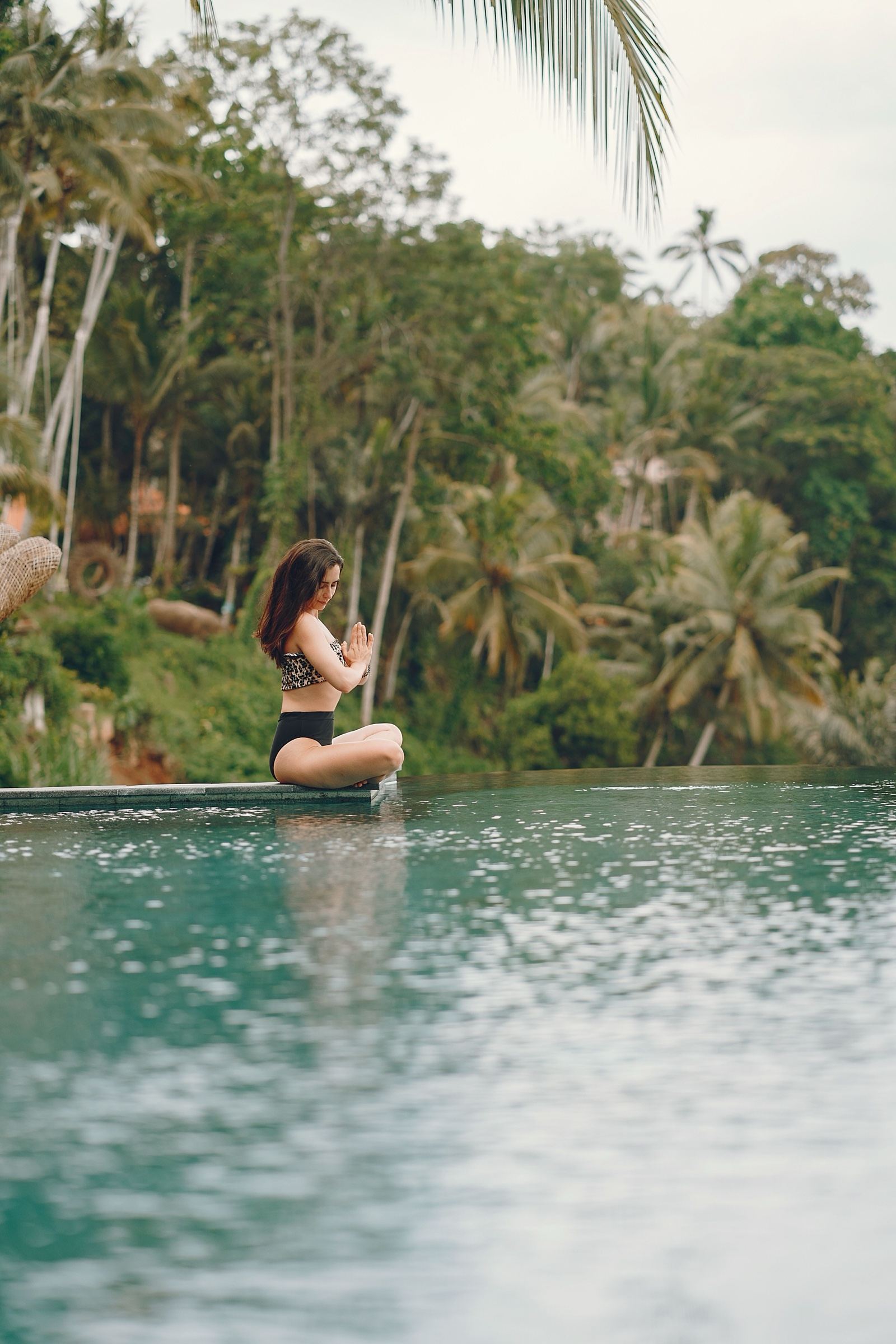 A woman sitting cross-legged with her hands together at the edge of a pool among palm trees in Indonesia
